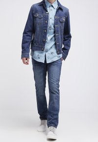 Persoon die loopt in een blauwe denimjack, een bedrukte lichtblauwe shirt, blauwe jeans en witte sneakers tegen een effen achtergrond.