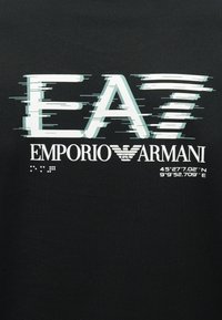 EA7 logó mozgási vonalakkal az "EMPORIO ARMANI" szöveg és a márka sas szimbóluma felett, 45°27'7,02"É 9°9'52,709"K koordinátákkal fekete háttéren.