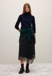 Pull à col roulé bleu marine, châle vert noué à la taille, jupe à carreaux avec des nuances de noir et vert, ourlet à franges, associé à des chaussures noires à lacets.