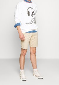 Sudadera blanca con un diseño de Snoopy en negro, superpuesta sobre una camisa de mezclilla azul. Unos pantalones cortos beige y zapatillas altas beige completan el conjunto.