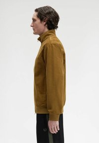 Uomo con capelli ricci che indossa una giacca in corduroy senape e pantaloni neri, in profilo su uno sfondo bianco uniforme.