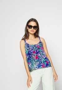 Top de flores com um fundo azul marinho, apresentando padrões vibrantes em rosa e verde, alças finas e um corte solto. Usado com calças de tom claro.