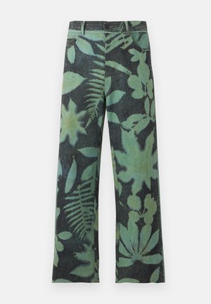 Νέος άνδρας που φοράει μαύρο καπέλο, λευκό κοντομάνικο πουκάμισο με διακριτικό σχέδιο, φαρδιά πράσινα και μαύρα φλοράλ παντελόνια και μαύρα και λευκά παπούτσια.