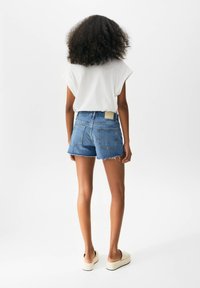 Denim shorts dengan tepi yang koyak, berwarna biru muda, dua saku belakang, dan label cokelat muda di bagian pinggang. Dipadukan dengan kaos putih.