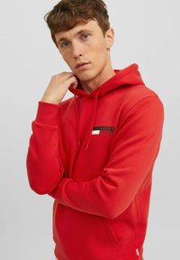 Sudadera roja hecha de un tejido con una textura suave, que presenta mangas largas, capucha con cordón y un pequeño parche con el logo en el pecho.