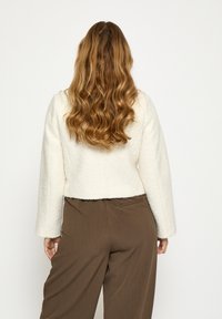 Kremiškai spalvos trumpa striukė su tekstūruota apdaila ir plačiomis rankovėmis, derinama su rudos spalvos platėjanciais kelnais. Nugara matyti švelnios medžiagos detalę.