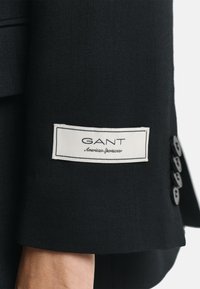 Zwarte jacksleeve met een wit label met "GANT" en "American Sportswear." Vijf zwarte knopen langs de manchet. Geweven stof.