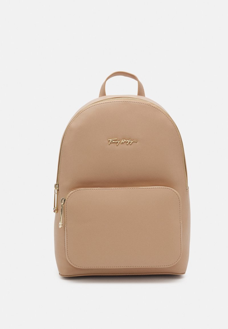Beige backpack featuring a rounded shape, smooth synthetic material, a front zippered pocket, and gold-tone hardware accents.
