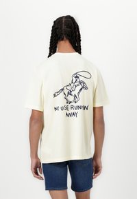 T-shirt en coton jaune clair avec un graphique au dos repr�ésentant un cavalier sur un cheval, et le texte "PAS D'UTILITÉ À FUIR" en lettres bleu foncé.