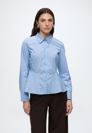 Woman wearing a blue striped button-up shirt with a peplum hem and dark pants, standing against a plain white background.