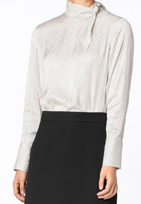 Vit blus med diagonala svarta ränder, hög hals och detaljer med knytblus. Långa ärmar med knäppte manschetter. Matchas med en svart kjol.