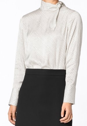 Blusa blanca con rayas diagonales negras, cuello alto y detalle de lazo. Mangas largas con puños abotonados. Combinada con una falda negra.