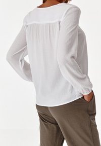 Blusa bianca a maniche lunghe con un motivo testurizzato, scollo arrotondato, polsini elasticizzati e dettaglio arricciato sul retro, abbinata a pantaloni verde oliva.