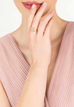 Gold ring featuring a 'W' design with a small stone accent, worn on a hand, against a background of soft pink fabric with pleats.