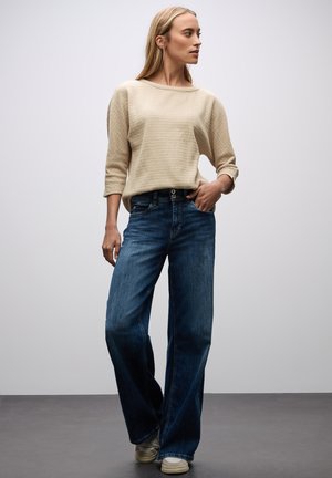 Beige gebreide trui met wijde nek, gecombineerd met losvallende diepblauwe jeans met twee knoopsluitingen en subtiele vervaging.