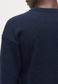 Pull en maille bleu marine avec des motifs texturés et un col côtelé. Le tissu présente un design côtelé classique avec un léger éclat.