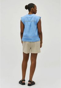 Lichtblauwe mouwloze top met een gestructureerde kantafwerking, gecombineerd met beige shorts. De top heeft een plooitje aan de achterkant en een knoopdetail. Zwarte sandalen.