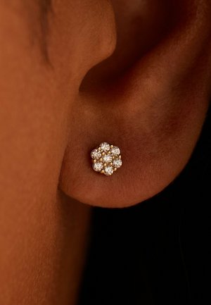 Gouden oorring in bloemvorm met zes ronde, heldere stenen in een cluster, met een gepolijste textuur tegen een bruin oor.