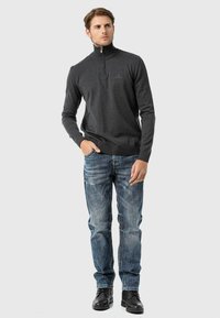 Grauer Reißverschluss-Pullover aus weichem Material, kombiniert mit faded blauen Jeans. Auffällige Details sind die gerippten Bündchen und der Stehkragen. Schwarze Stiefel runden den Look ab.