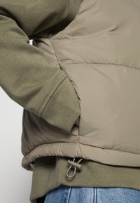 Puffervest i lysebeige over en grøn sweatshirt. Har en sidelomme og justerbar kant med snor og lås. Blødt, tekstureret stof.