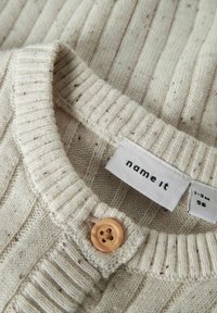 Pull en tricot beige clair avec une texture côtelée, doté d'un bouton en bois et d'une étiquette portant la mention "name it", avec de petites taches réparties sur le tissu.