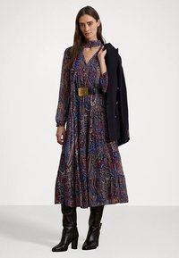 Pitkä, kuvioitu mekko sinisen, punaisen ja keltaisen paisley-kuvion kanssa, korkea kaulus ja pitkät hihat, yhdistettynä mustaan vyöhön ja polven ylle ulottuviin mustiin saappaisiin.