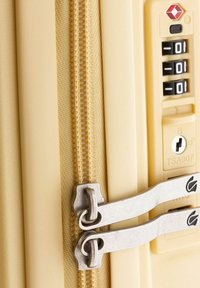 Equipaje color crema con un exterior texturizado, que presenta cremalleras metálicas dobles, una cerradura combinada y un emblema de seguridad de la TSA.