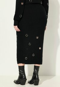 Gonna midi nera a maglia con grandi occhielli metallici. Il design presenta una silhouette aderente, una texture a coste e spacchi in fondo per la libertà di movimento.