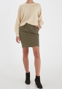 Žena oblečená v béžovém pleteném svetru, olivově zelené úzké sukni s kapsami a černých semišových kotníkových botách stojí na bílém pozadí.