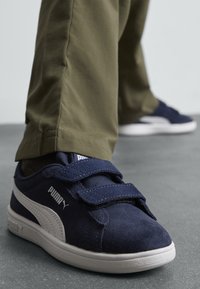 Baskets en daim bleu marine avec des bandes Velcro et des accents blancs. Semelle en caoutchouc et tige en textile, associées à un pantalon kaki.