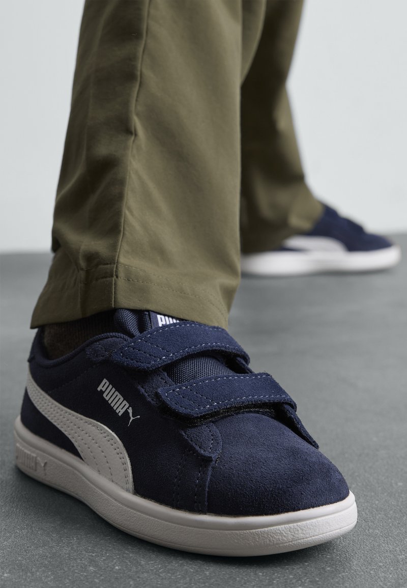 Baskets en daim bleu marine avec des bandes Velcro et des accents blancs. Semelle en caoutchouc et tige en textile, associées à un pantalon kaki.