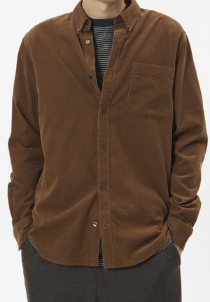 Hombre vistiendo una camisa marrón de pana con botones sobre una camiseta de rayas blancas y negras, con las manos en los bolsillos de pantalones gris oscuro.