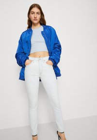 Kék selyem bomber dzseki egy világoskék crop top fölött, párosítva koptatott fehér skinny farmernadrággal és fekete magassarkú cipőkkel.