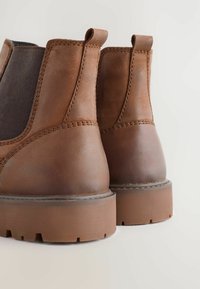 Ein Paar braune Chelsea-Stiefel aus Leder mit dicken Gummisohlen und elastischen Seitenpaneelen, von hinten auf einem weißen Hintergrund gezeigt.