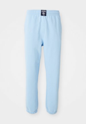 Lys blå joggebukser med elastisk midje og elastiske mansjetter, med et svart merke med hvit Lacoste Paris-logo på bakmidjen.