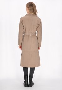 Lang beige trenchcoat lavet af blødt materiale, med et bælte i taljen, slagkrave og sideslids, parret med sorte ankelstøvler.