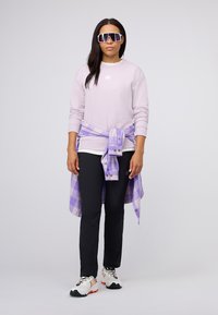 Lichtpaars sweatshirt, zwarte broek, witte geblokte flanellen blouse om de taille geknoopt, en witte sportieve schoenen met kleurrijke accenten. Grote zonnebril.