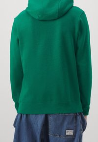 Grön hoodie av mjukt tyg, med huva och ribbade muddar. Kombinerad med lösa jeansbyxor med synlig varumärkeslapp.