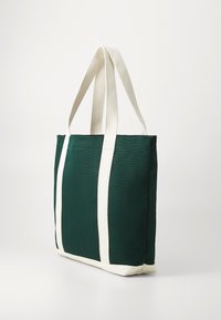 Grüne Canvas-Shopper-Tasche mit weißen Trägern und Boden, die aufrecht vor einem schlichten hellen Hintergrund steht.