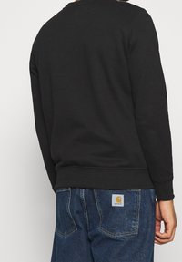 Felpa nera con scollo rotondo e polsini a coste, abbinata a jeans blu con una patch del logo Carhartt sulla tasca posteriore.