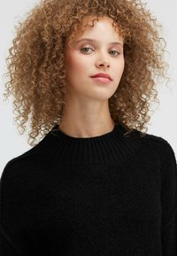 Frau mit lockigem, hellbraunem Haar, die einen schwarzen strukturierten Pullover trägt und leicht nach oben blickt, vor einem schlichten weißen Hintergrund.