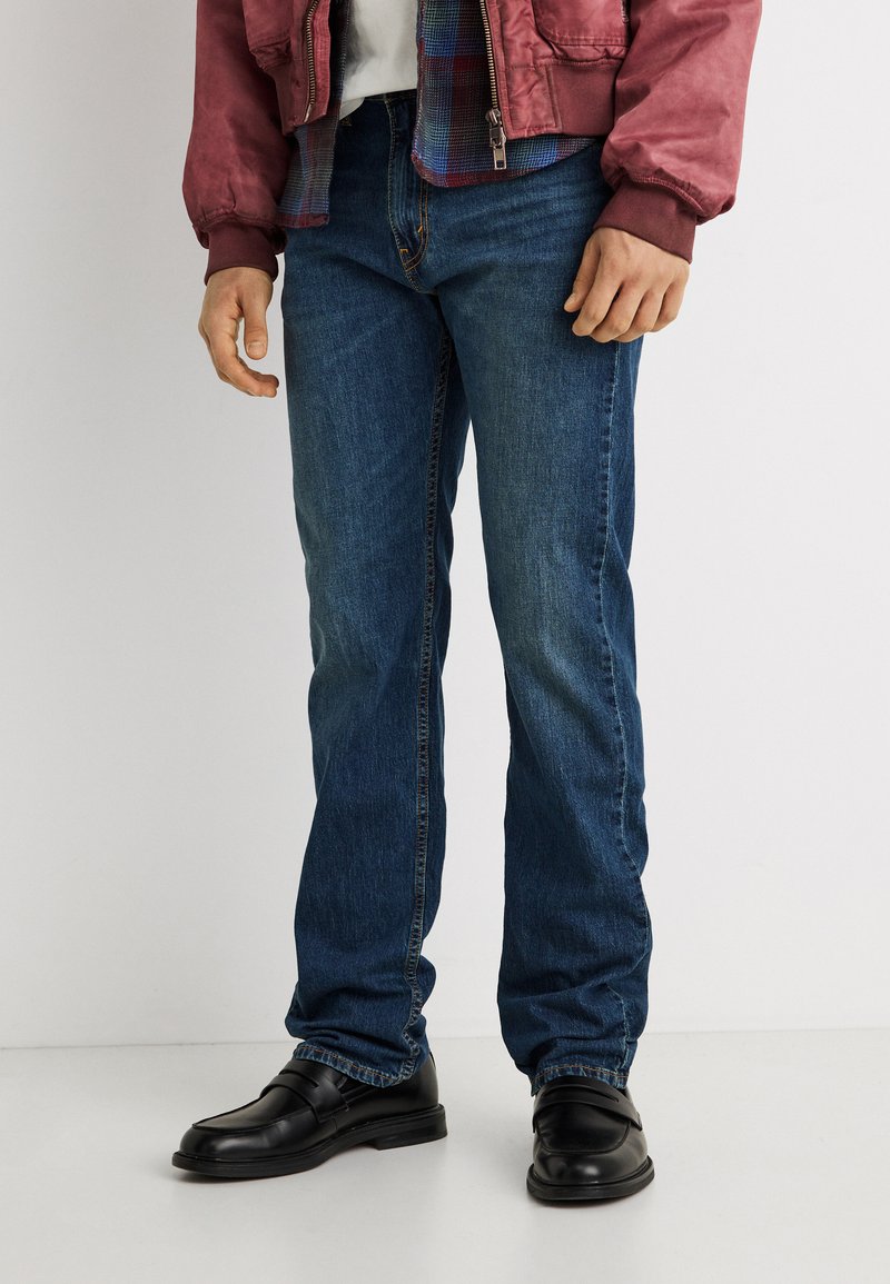 Άνδρας που φοράει σκουρό μπλε τζιν με ίσια γραμμή, μαύρα δερμάτινα μοκασίνια, κόκκινο μπουφάν και καρό πουκάμισο φορεμένο κάτω από το μπουφάν.