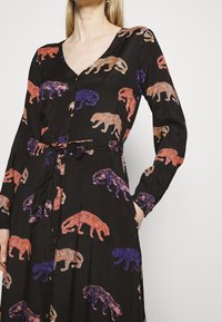 Svart klänning med ett upprepat mönster av färgglada leoparder i orange, lila och brunt. Designen inkluderar en V-ringning och en knytning i midjan.