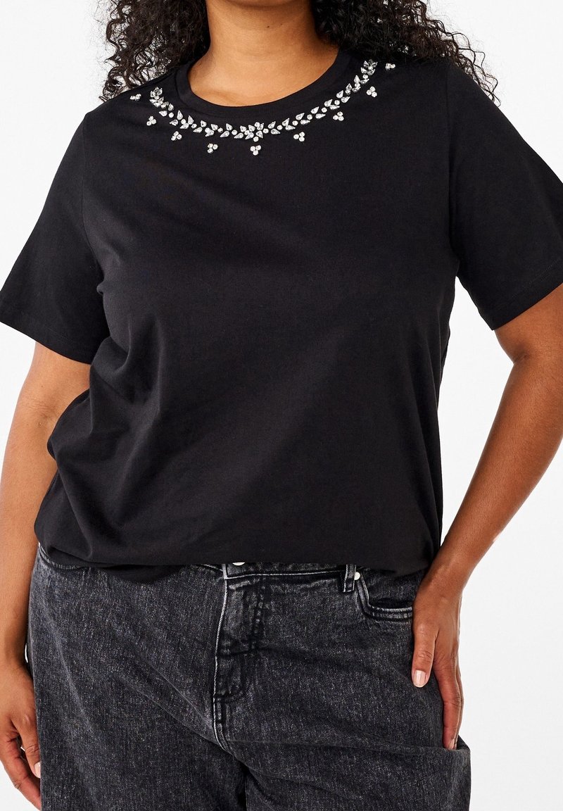 Femme portant un t-shirt noir à manches courtes avec un motif floral en strass près de l'encolure et un jean denim gris foncé.
