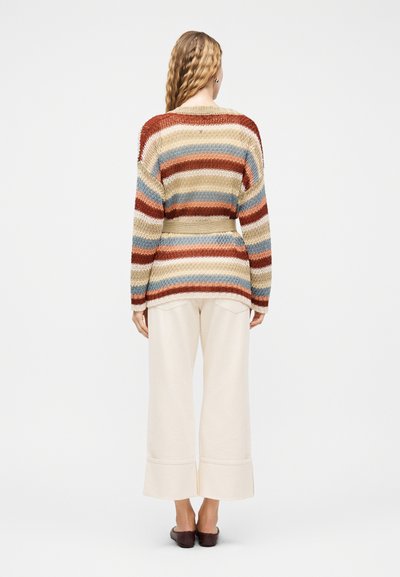 Γυναίκα με κυματιστά μαλλιά που φοράει ριγέ πλεκτό πουλόβερ σε γήινες αποχρώσεις, παντελόνι με φαρδιά πόδια σε κρεμ χρώμα και καφέ επίπεδα παπούτσια, όρθια με πλάτη γυρισμένη.