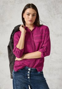 Camicia fucsia con bottoni, decorata con motivi geometrici neri, maniche lunghe arrotolate, abbinata a jeans in denim blu con bottoni bianchi.