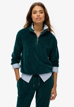 Teal fleece pullover met een halve ritssluiting en een kraag, gedragen over een lichtblauw gestreept shirt. Zachte textuur, relaxte pasvorm en opvallende stiksels.