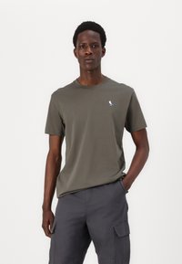 Tricou din bumbac de culoare verde măsliniu, cu guler rotund, mâneci scurte și un mic logo cu o pasăre albastră pe piept. Purtați cu pantaloni cargo gri.