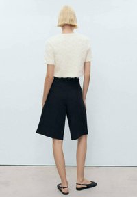 Vit texturerad kortärmad topp matchad med svarta knälånga skräddarsydda shorts. Modellen bär svarta platta skor. Enkelt design och neutrala färger.