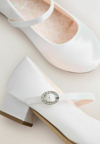 Białe lakierowane buty typu Mary Jane z okrągłym czubkiem, regulowanym paskiem i ozdobną owalną klamrą zdobioną przezroczystymi cyrkoniami.